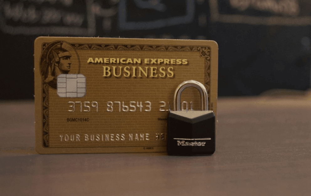 A brown American Express credit card stands on a table, and a small black padlock stands in front of it.