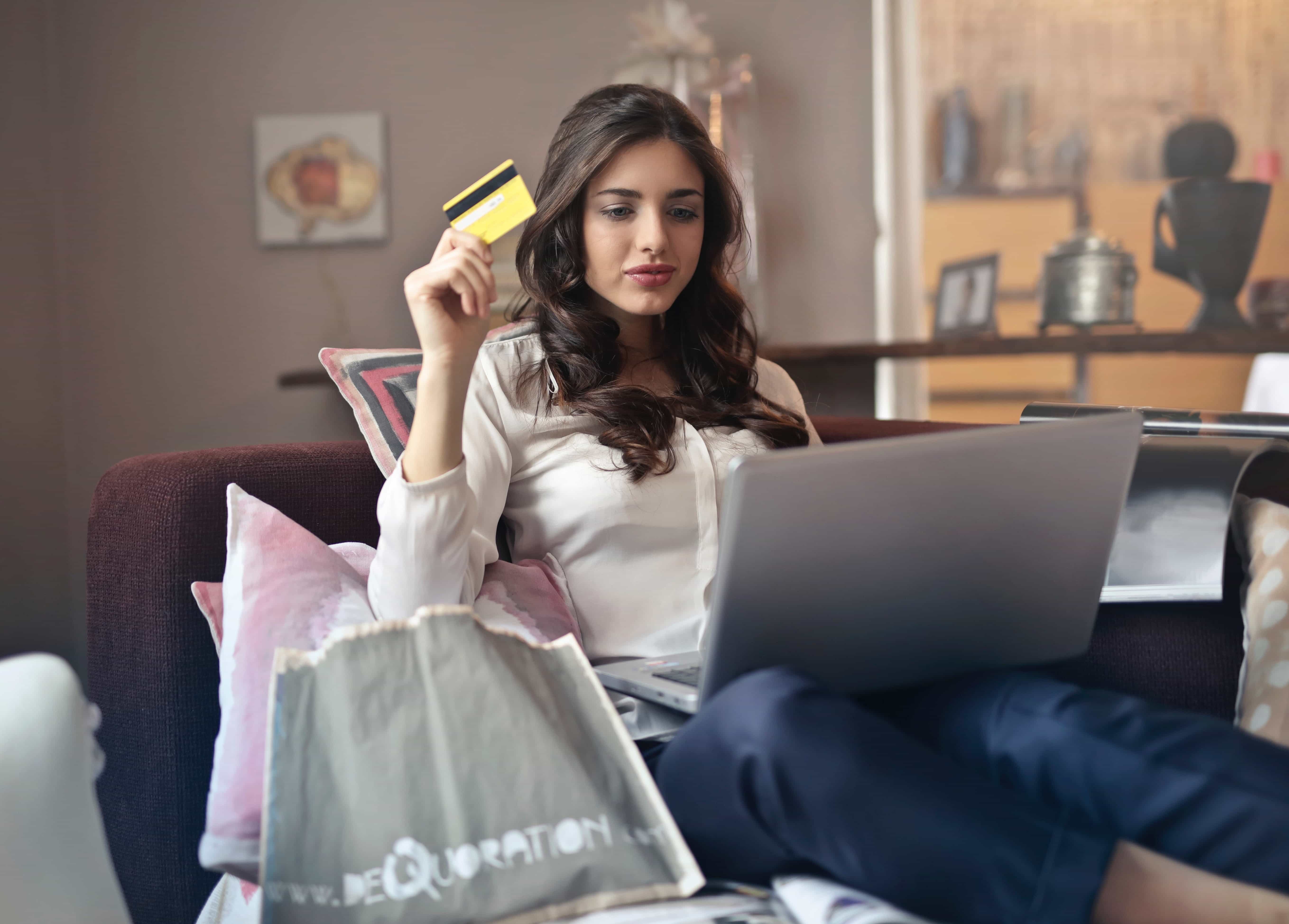 A white woman holds up a credit card while sitting with a silver laptop on her lap.