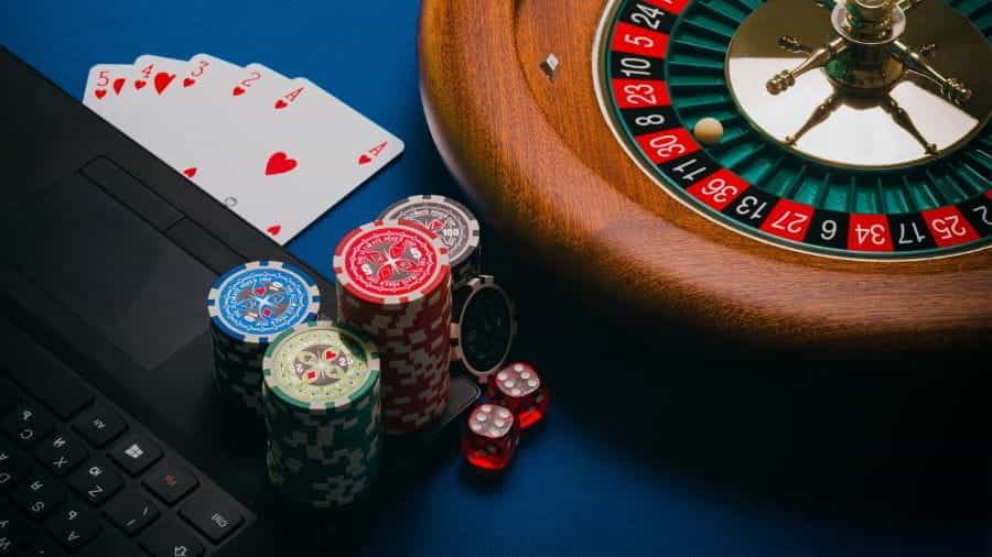 A roulette wheel, playing cards, dice and poker chips next to a laptop.