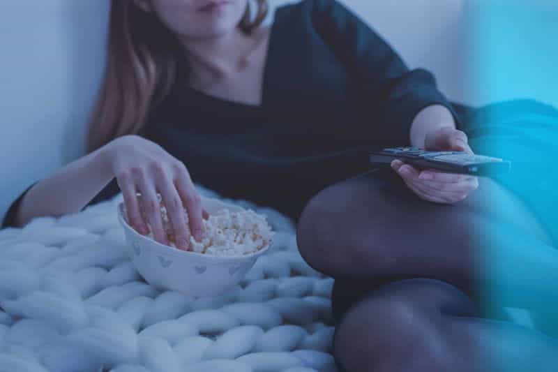 A woman sitting on a sofa watching the television with popcorn.
