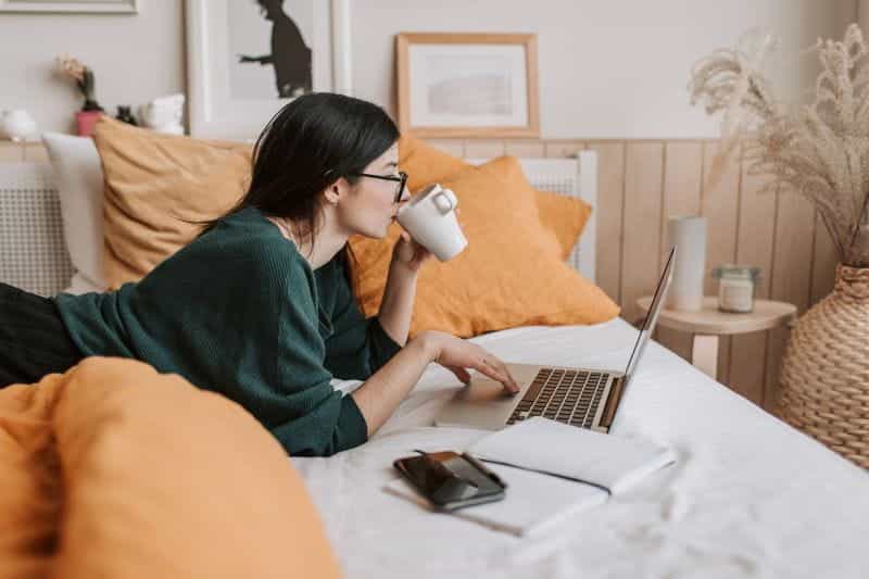 A woman drinking coffee and using a laptop on the bed.