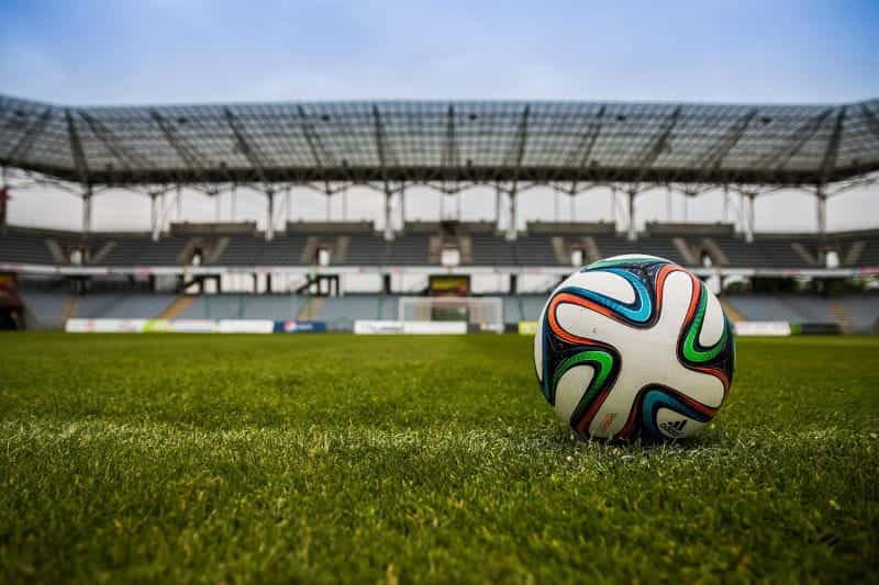 A soccer ball resting on the grass of a professional soccer pitch in a large and empty stadium.