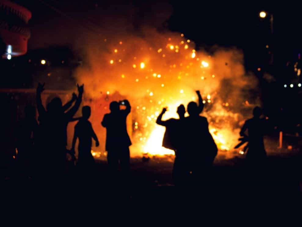 Silhouetted people raise their arms in front of a large fire.