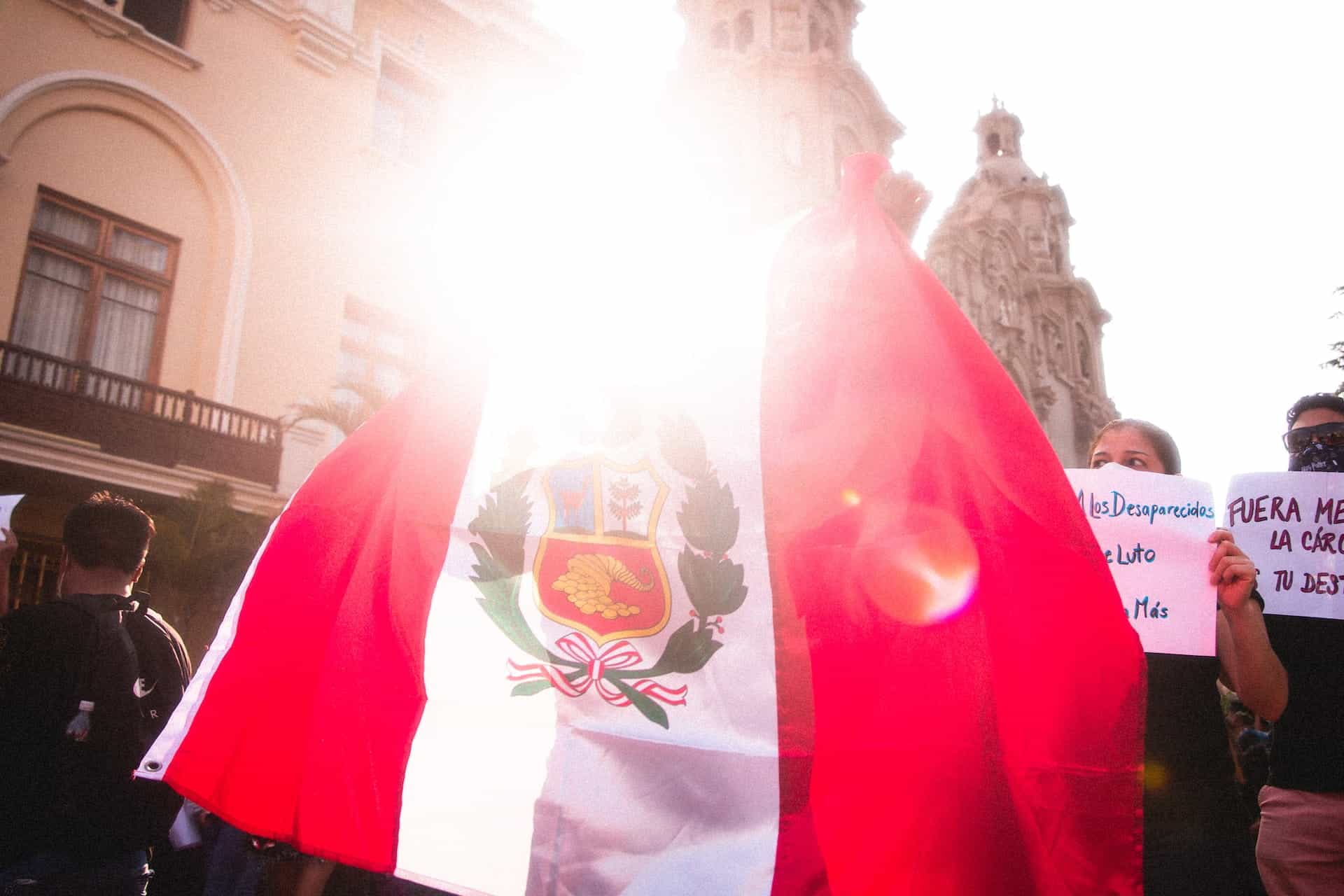The Peruvian flag waves in front of a marching crowd, brightened by sunlight behind it.