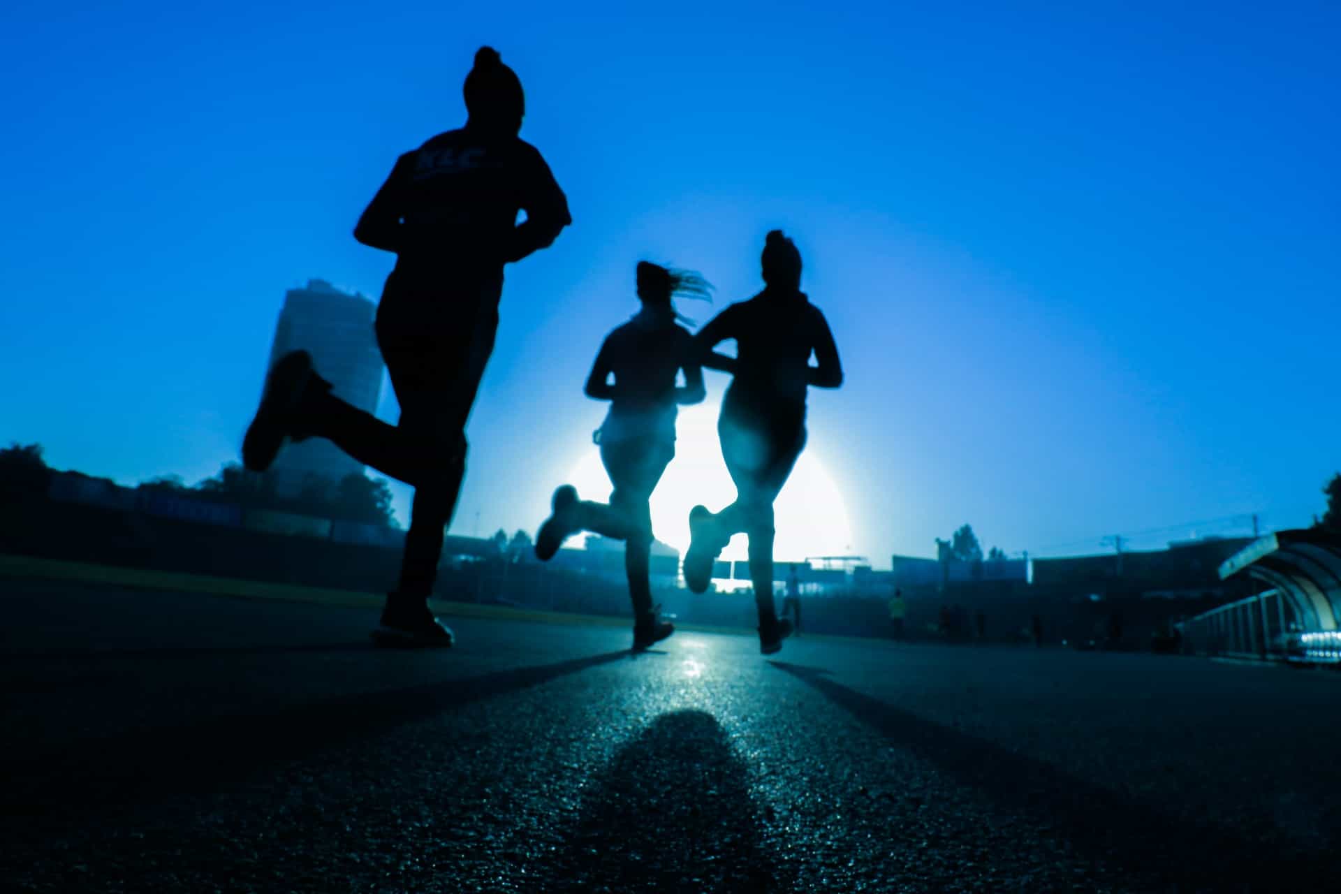 Three runners jogging together outside on an outdoor track and field track, with a large building towering above them in the background.