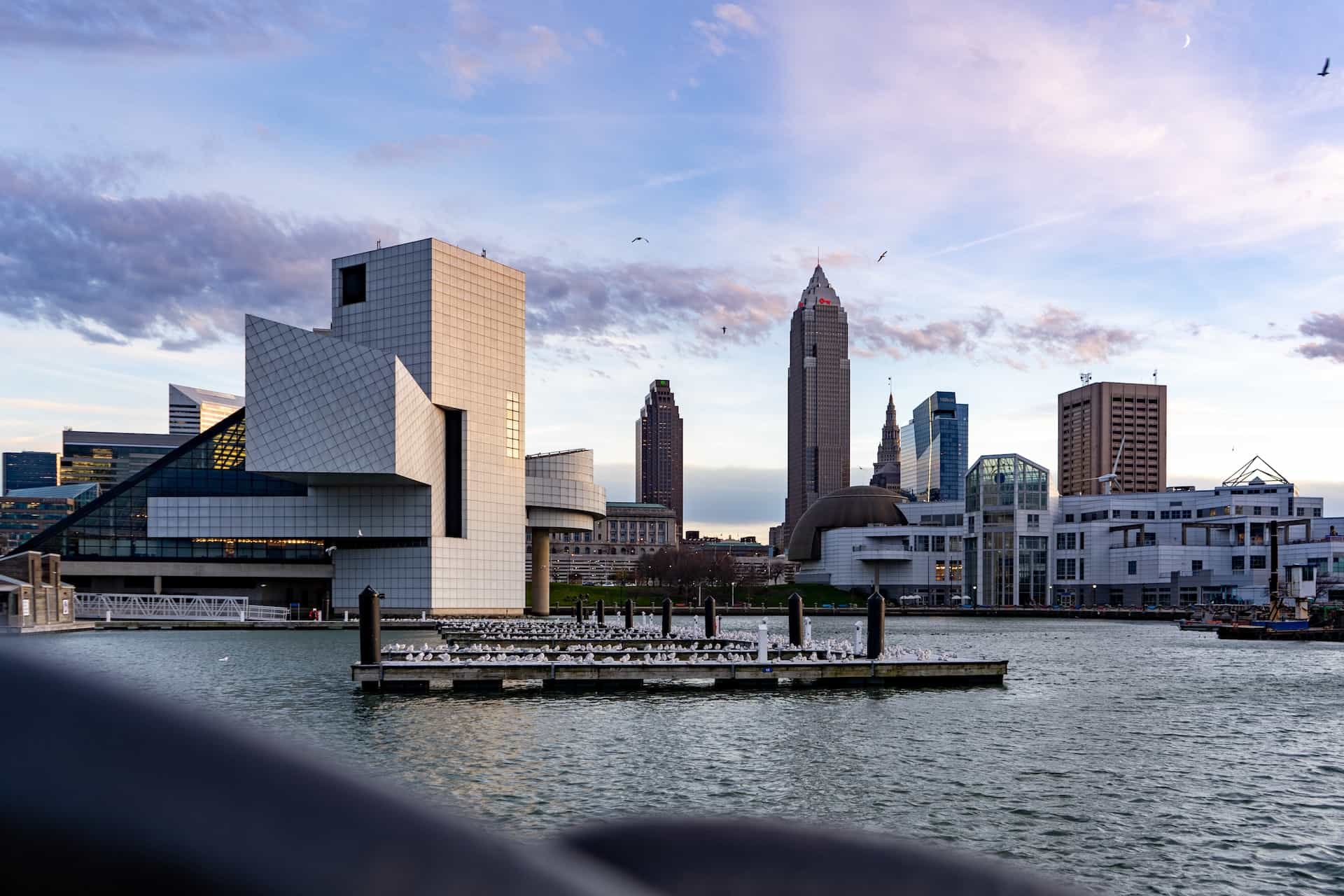 A view of Cleveland’s downtown district from its harbor, with the world-famous Rock and Roll Hall of Fame situated to the left, in front of tall buildings and skyscrapers in the background.